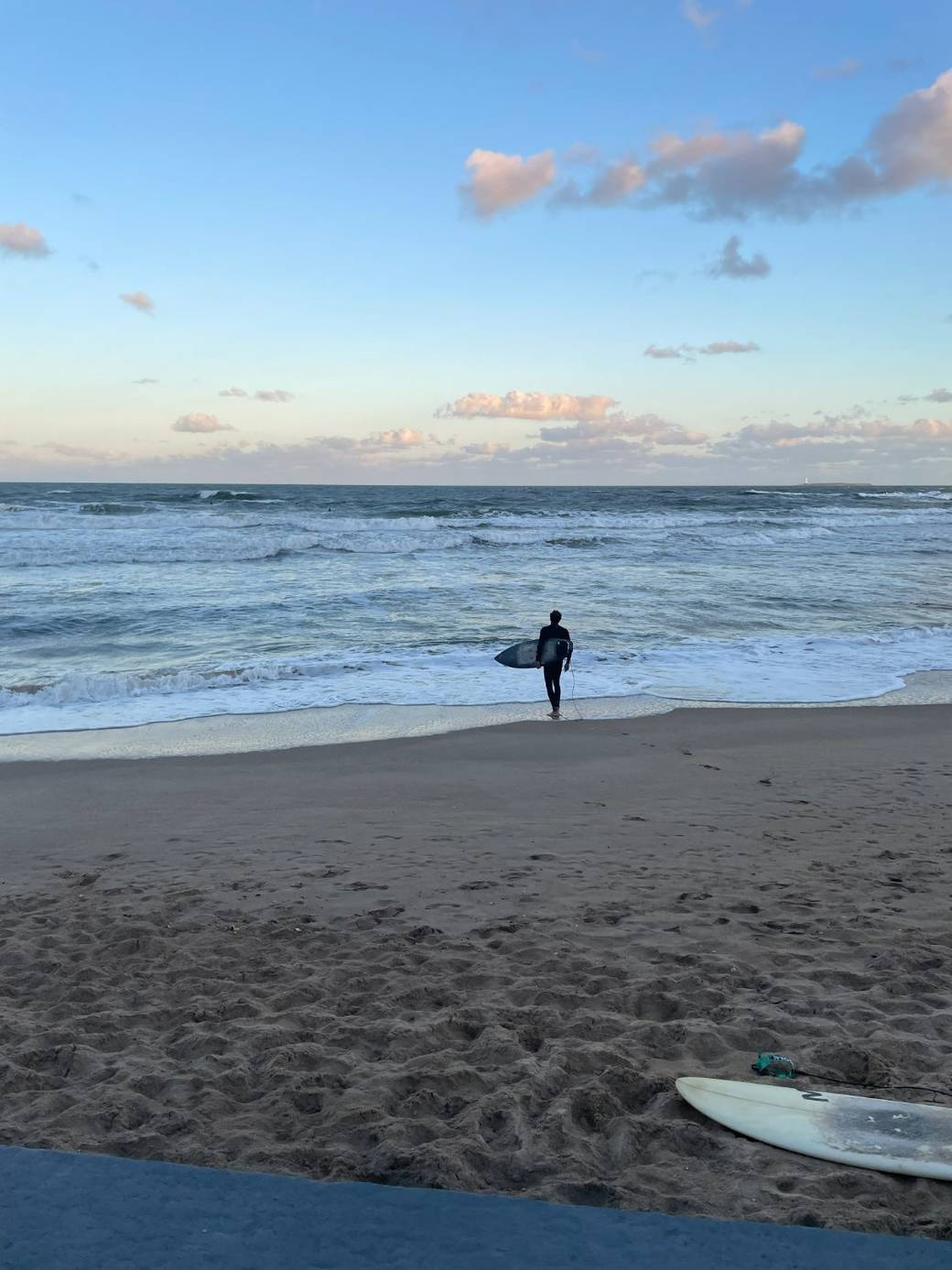 Una persona caminando sola por la orilla del mar al atardecer, con la tabla de surf